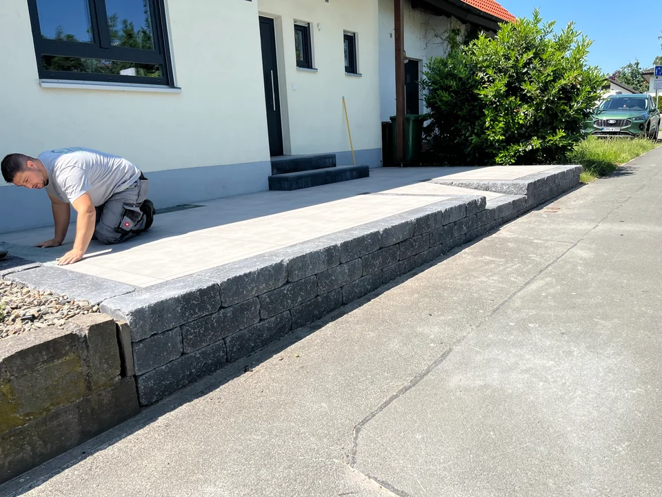 Erhöhte Vorfläche aus hellen Platten vor einem Wohnhaus, eingefasst von einer Mauer aus dunkelgrauen Blocksteinen, ein Handwerker bei der Arbeit