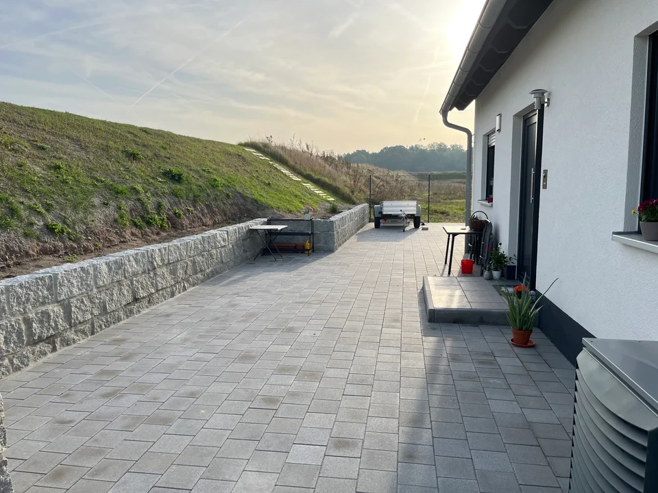 Gepflasterte Fläche entlang eines Wohnhauses mit Natursteinmauer vor einer begrünten Böschung
